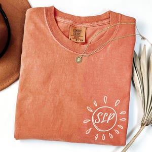 May include: Coral-colored Comfort Colors t-shirt with a white sun graphic and the letters "SLP" inside. A gold necklace with a pendant rests on the shirt. A brown hat and dried plant are in the background.