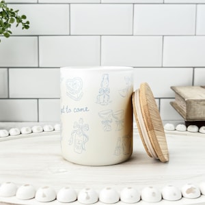 May include: A cream-colored ceramic jar with a bamboo lid, decorated with light blue illustrations and the words "yet to come." The jar is on a white tray with white decorative beads, suggesting a kitchen or home decor theme.