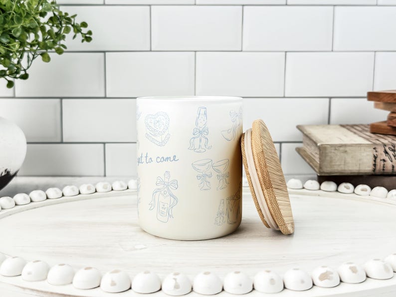 May include: A cream-colored ceramic jar with a bamboo lid, decorated with light blue illustrations and the words "yet to come." The jar is on a white tray with white decorative beads, suggesting a kitchen or home decor theme.