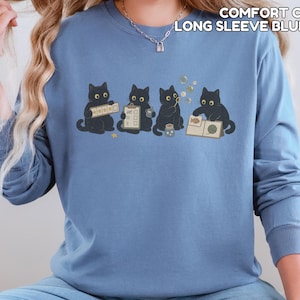 May include: A blue jean long-sleeve crewneck sweatshirt featuring a graphic of four black cats engaged in various activities, including holding a clipboard and blowing bubbles. The text COMFORT COLORS LONG SLEEVE BLUE JEAN is printed in the upper right corner.