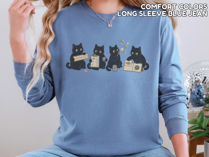 May include: A blue jean long-sleeve crewneck sweatshirt featuring a graphic of four black cats engaged in various activities, including holding a clipboard and blowing bubbles. The text COMFORT COLORS LONG SLEEVE BLUE JEAN is printed in the upper right corner.
