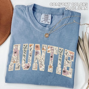 Puede incluir: Una camiseta azul vaquero Comfort Colors con la palabra "AUNTIE" en letras grandes y florales. La camiseta tiene cuello redondo y mangas cortas. Un collar dorado con un colgante est&aacute; drapeado sobre la camiseta.