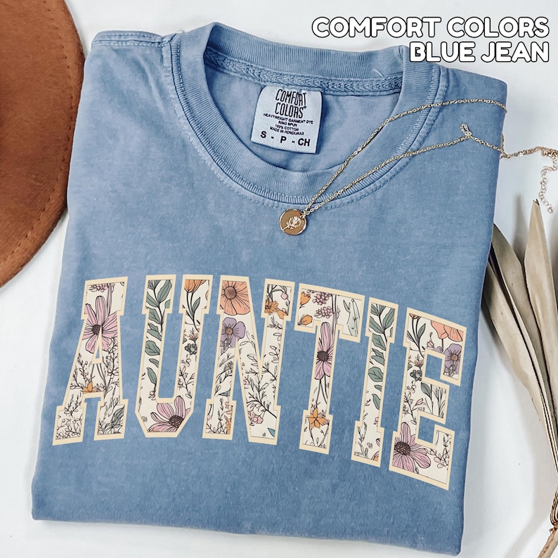 Puede incluir: Una camiseta azul vaquero Comfort Colors con la palabra "AUNTIE" en letras grandes y florales. La camiseta tiene cuello redondo y mangas cortas. Un collar dorado con un colgante est&aacute; drapeado sobre la camiseta.
