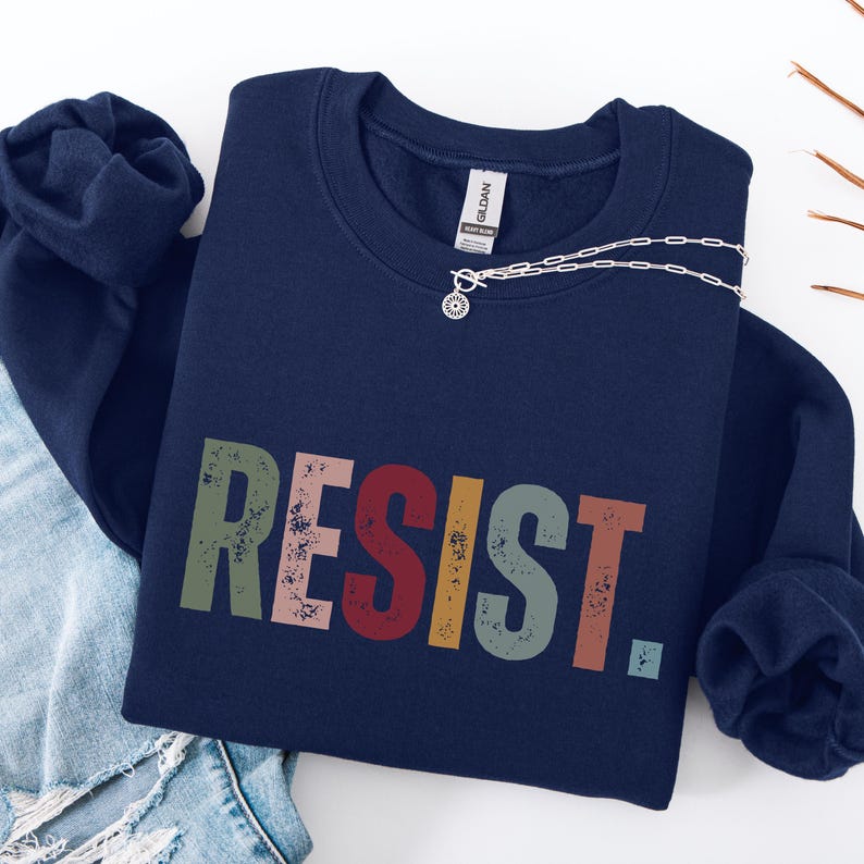 Pode incluir: Uma camisa de moletom azul marinho com a palavra "RESIST" impressa em letras coloridas e desgastadas. As letras s&atilde;o em tons de verde, rosa, vermelho e amarelo.
