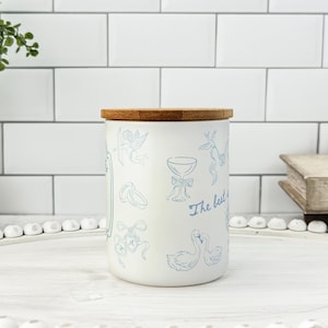 May include: A white ceramic jar with a bamboo lid, decorated with light blue illustrations of wedding-themed elements. The jar has the text "The best is yet to come" in a script font.