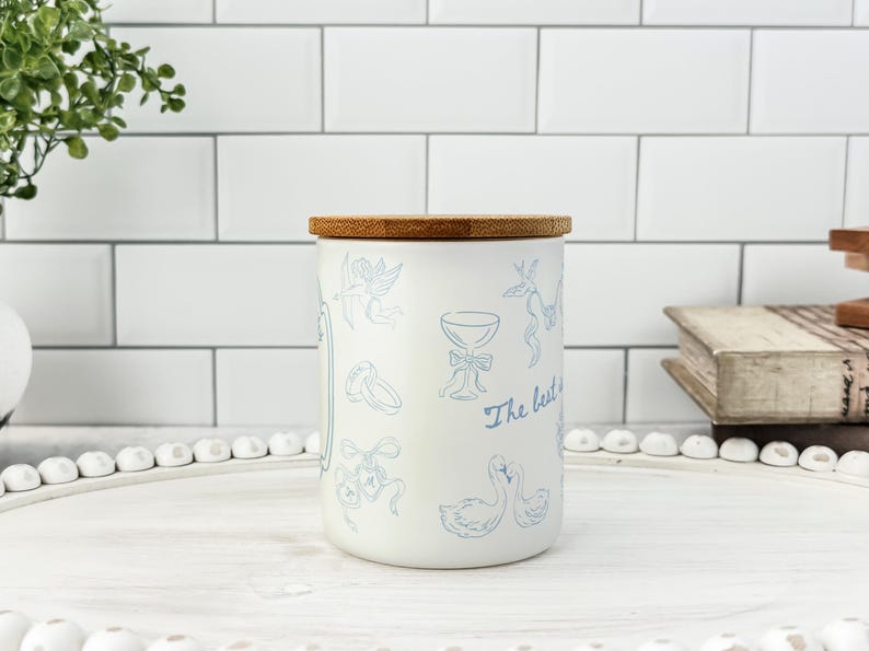 May include: A white ceramic jar with a bamboo lid, decorated with light blue illustrations of wedding-themed elements. The jar has the text "The best is yet to come" in a script font.