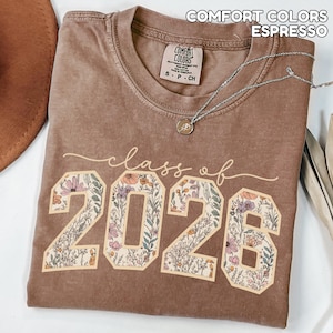 May include: Espresso-colored t-shirt with the text "class of 2026" in a cursive font. The numbers "2026" are filled with a floral design. The shirt has a crew neck and a small tag. A silver necklace with a pendant is draped across the shirt.
