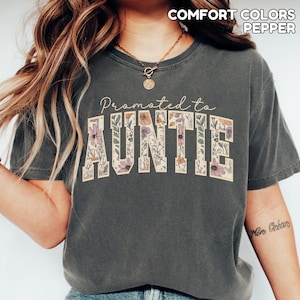 Puede incluir: Camiseta gris oscuro con el texto "Promoted to Auntie" en un dise&ntilde;o floral. La palabra "AUNTIE" est&aacute; en un estilo de letras en bloque, llena de varias flores y hojas. La camiseta tambi&eacute;n tiene las palabras "COMFORT COLORS PEPPER" impresas en la parte superior.