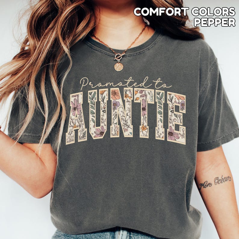 Puede incluir: Camiseta gris oscuro con el texto "Promoted to Auntie" en un dise&ntilde;o floral. La palabra "AUNTIE" est&aacute; en un estilo de letras en bloque, llena de varias flores y hojas. La camiseta tambi&eacute;n tiene las palabras "COMFORT COLORS PEPPER" impresas en la parte superior.