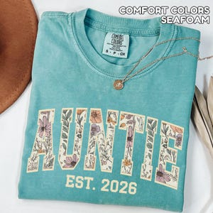 Puede incluir: Camiseta verde mar con la palabra "AUNTIE" en un dise&ntilde;o floral y el texto "EST. 2026". La camiseta tiene cuello redondo y una peque&ntilde;a etiqueta en el cuello. Un collar dorado con un colgante est&aacute; drapeado sobre la camiseta.