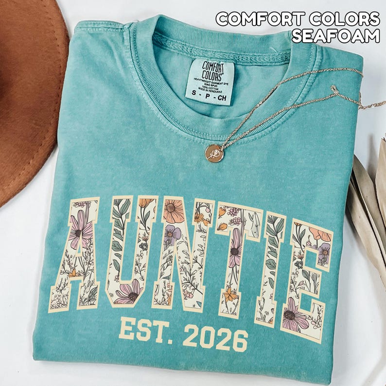 Puede incluir: Camiseta verde mar con la palabra "AUNTIE" en un dise&ntilde;o floral y el texto "EST. 2026". La camiseta tiene cuello redondo y una peque&ntilde;a etiqueta en el cuello. Un collar dorado con un colgante est&aacute; drapeado sobre la camiseta.
