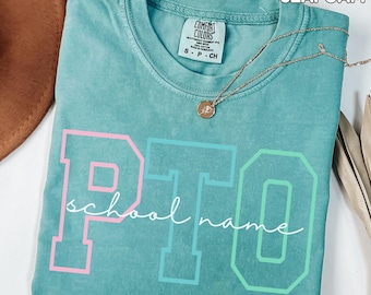 May include: Seafoam green t-shirt with the text "PTO school name" in pink and green block letters. The shirt has a crew neck and short sleeves. A gold necklace with a pendant is draped across the shirt.