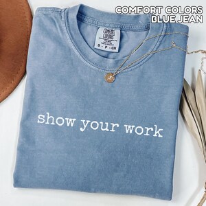 K&ouml;nnte beinhalten: Ein blaues Comfort Colors T-Shirt im Jeans-Look mit dem Schriftzug "show your work" in wei&szlig;en Buchstaben. Eine goldene Halskette mit Anh&auml;nger liegt auf dem Shirt. Das Shirt ist gefaltet und hat ein kleines Etikett am Ausschnitt.