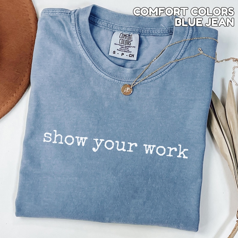 K&ouml;nnte beinhalten: Ein blaues Comfort Colors T-Shirt im Jeans-Look mit dem Schriftzug "show your work" in wei&szlig;en Buchstaben. Eine goldene Halskette mit Anh&auml;nger liegt auf dem Shirt. Das Shirt ist gefaltet und hat ein kleines Etikett am Ausschnitt.