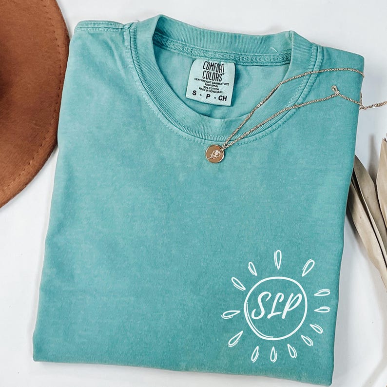 May include: A teal Comfort Colors t-shirt with a white sun graphic and the letters "SLP" inside. A gold necklace with a pendant is draped across the shirt. The shirt is size small.