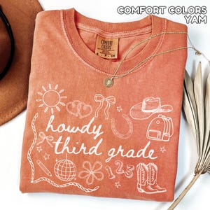 May include: A coral-coloured t-shirt with the words "howdy third grade" and various white illustrations, including a sun, hearts, a bow, a horseshoe, a cowboy hat, a backpack, a pencil, a disco ball, a flower, the numbers 123, and cowboy boots. A gold necklace is draped across the shirt.
