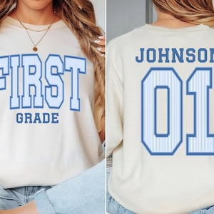 May include: A cream-colored t-shirt featuring blue text with a white pinstripe pattern. The front displays the words FIRST GRADE in a collegiate font. The back shows the name JOHNSON above the large jersey-style number 01 in the same blue font.