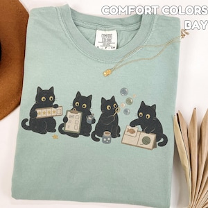May include: A sage green t-shirt featuring a graphic of four black cats engaged in various activities, including holding a five-star rating card, a checklist, blowing bubbles, and looking at a book. The shirt has a Comfort Colors label.