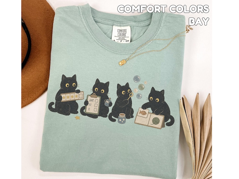 May include: A sage green t-shirt featuring a graphic of four black cats engaged in various activities, including holding a five-star rating card, a checklist, blowing bubbles, and looking at a book. The shirt has a Comfort Colors label.