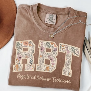 May include: A brown t-shirt with a floral design spelling out "RBT" and the text "Registered Behaviour Technician".