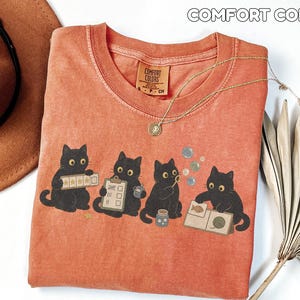 May include: A terracotta-colored t-shirt featuring a graphic of four black cats engaged in various activities, including holding a five-star rating card, a clipboard, a bubble wand, and a book. The shirt has a Comfort Colors label.