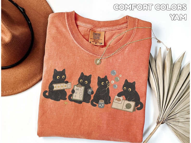 May include: A terracotta-colored t-shirt featuring a graphic of four black cats engaged in various activities, including holding a five-star rating card, a clipboard, a bubble wand, and a book. The shirt has a Comfort Colors label.