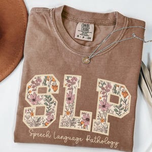 May include: A brown t-shirt with a floral design spelling out "SLP" and the text "Speech Language Pathology" below.