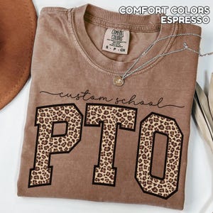 May include: Espresso-colored t-shirt with the text "custom school PTO" in black script and block letters. The letters "PTO" are filled with a leopard print pattern. The shirt has a crew neck and short sleeves. A silver necklace with a pendant is draped across the shirt.