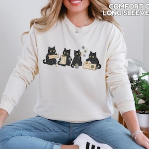 May include: An ivory long-sleeve sweatshirt featuring a graphic of four black cats engaged in various activities, including holding a sign, a clipboard, blowing bubbles, and reading a book. The text COMFORT COLORS LONG SLEEVE IVORY is visible.