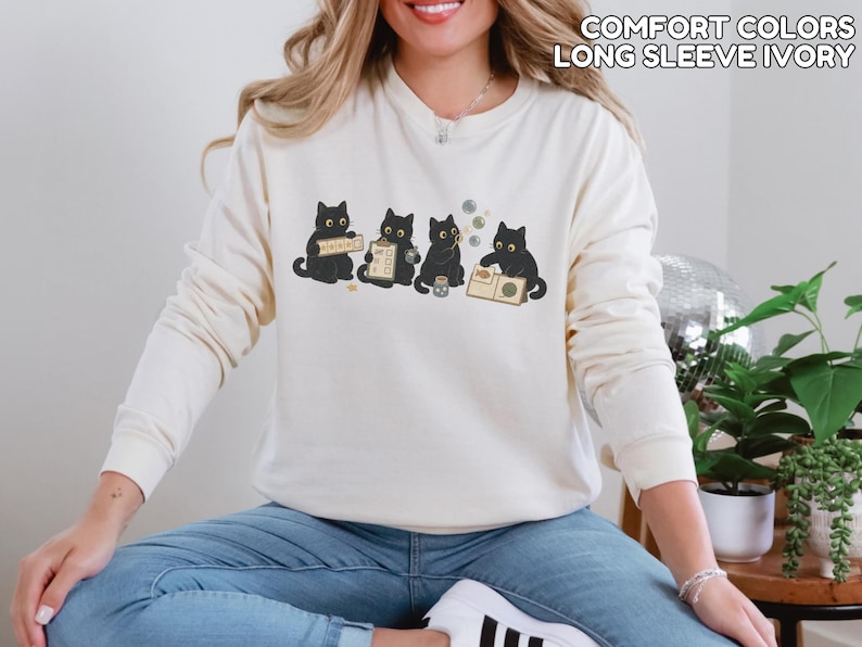 May include: An ivory long-sleeve sweatshirt featuring a graphic of four black cats engaged in various activities, including holding a sign, a clipboard, blowing bubbles, and reading a book. The text COMFORT COLORS LONG SLEEVE IVORY is visible.