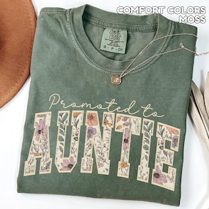 Puede incluir: Una camiseta verde musgo con las palabras "Promoted to AUNTIE" en un dise&ntilde;o floral. La camiseta tiene cuello redondo y mangas cortas. Un collar dorado con un colgante est&aacute; drapeado sobre la camiseta.