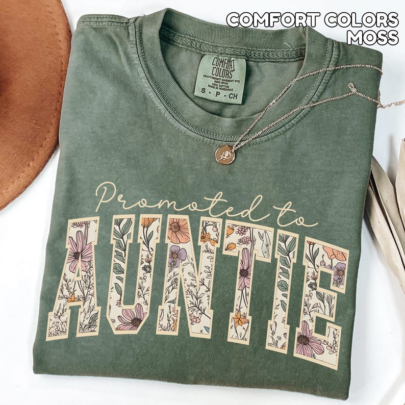 Puede incluir: Una camiseta verde musgo con las palabras "Promoted to AUNTIE" en un dise&ntilde;o floral. La camiseta tiene cuello redondo y mangas cortas. Un collar dorado con un colgante est&aacute; drapeado sobre la camiseta.