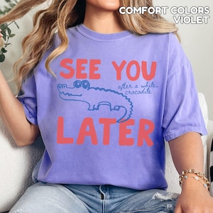 May include: A lilac-coloured t-shirt with the words "SEE YOU LATER" in coral-red block letters. A blue cartoon crocodile is between the words. The text "after a while, crocodile" is above the crocodile.