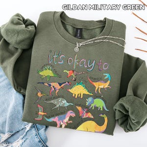 May include: A military green sweatshirt with the text "it's okay to" and colorful dinosaur illustrations. A silver necklace with a circular pendant is draped across the sweatshirt, which is folded next to a pair of blue jeans.