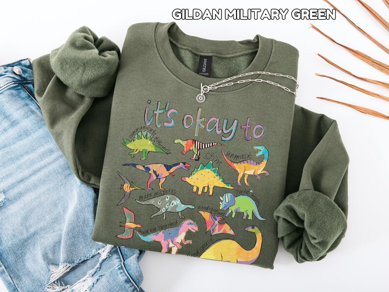 May include: A military green sweatshirt with the text "it's okay to" and colorful dinosaur illustrations. A silver necklace with a circular pendant is draped across the sweatshirt, which is folded next to a pair of blue jeans.