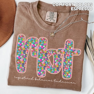 May include: Espresso-coloured t-shirt featuring the letters "RBT" in a floral design. The words "registered behaviour technician" are printed below the letters. The shirt also has the words "Comfort Colors" printed at the top.