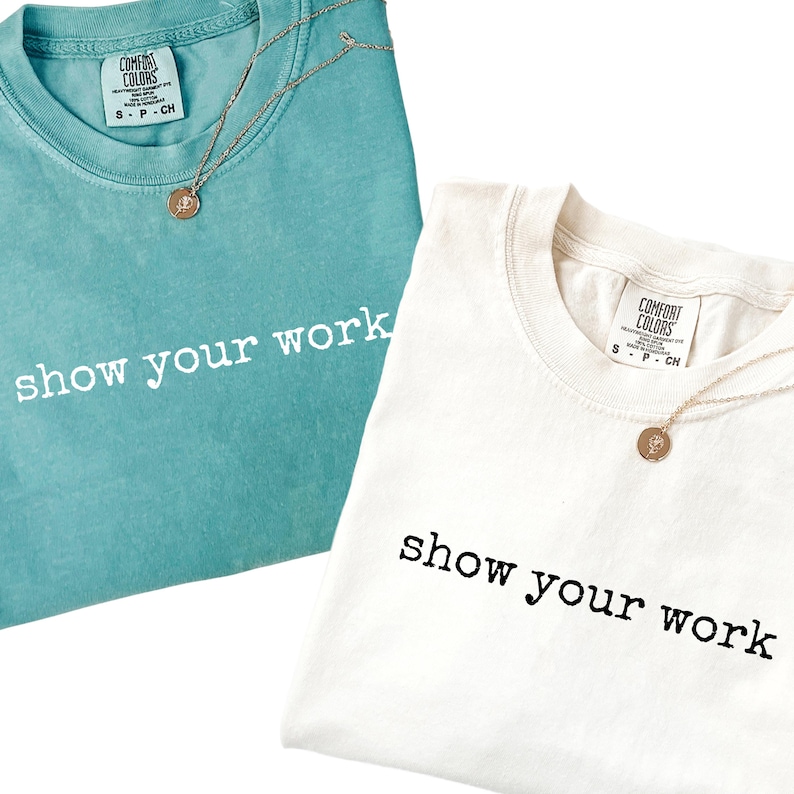 K&ouml;nnte beinhalten: Zwei T-Shirts, eines in T&uuml;rkis und eines in Off-White, mit dem Aufdruck "show your work" in Schwarz. Jedes Shirt hat ein Comfort Colors-Etikett und eine goldfarbene Halskette. Das t&uuml;rkisfarbene Shirt befindet sich links, das off-white Shirt rechts.