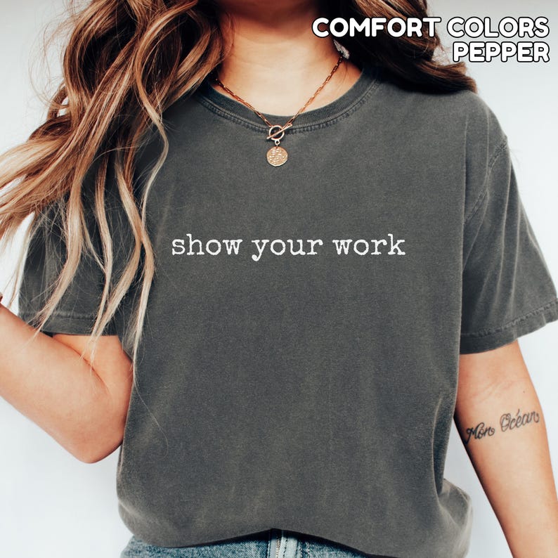 K&ouml;nnte beinhalten: Dunkelgraues T-Shirt mit dem wei&szlig;en Text "show your work". Das Shirt hat auch die Worte "COMFORT COLORS PEPPER" oben aufgedruckt. Das Shirt hat einen Rundhalsausschnitt und kurze &Auml;rmel.