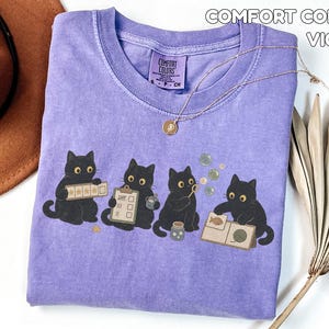 May include: A violet Comfort Colors t-shirt featuring a graphic of four black cats. The cats are shown interacting with various items including a star rating board, a checklist, a bubble wand, and a book with a fish and yarn illustration.