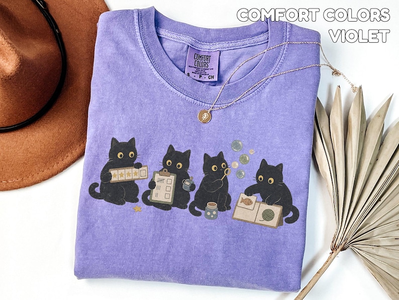 May include: A violet Comfort Colors t-shirt featuring a graphic of four black cats. The cats are shown interacting with various items including a star rating board, a checklist, a bubble wand, and a book with a fish and yarn illustration.