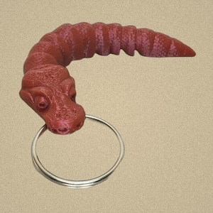 May include: A dark red, 3D-printed articulated snake keychain featuring a textured, segmented body that curves into a hook shape. The snake head is detailed with visible eyes and nostrils, and it holds a circular silver metal split ring.