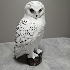 May include: A white snowy owl figurine with black speckled markings on its wings and back. The owl has dark eyes and a sharp beak, and it is perched on a textured, dark brown cylindrical base that resembles a tree stump.