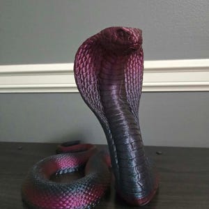 May include: A cobra figurine with a metallic finish that transitions from deep black to vibrant magenta. The snake is depicted in an upright, alert posture with its hood flared, showcasing detailed scale textures across its entire body.