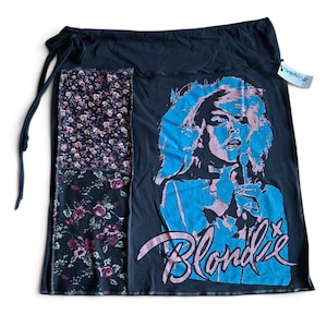 May include: A black skirt featuring a graphic print of a person singing into a microphone with the text Blondie in pink. The skirt includes a side panel with a floral pattern in shades of pink, white, and green, and a drawstring waist tie.