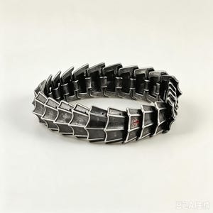 Op de afbeelding: Een zilverkleurige metalen armband met een gesegmenteerd, gearticuleerd ontwerp dat lijkt op overlappende pantserplaten. Het oppervlak heeft een verweerde, donkere afwerking met een klein, rood symbool gegraveerd op een van de metalen schakels.