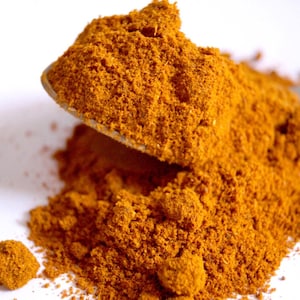 Puede incluir: Una cuchara de metal sostiene un montón de polvo de cúrcuma finamente molido de color naranja amarillento vibrante. Un montón más grande de la misma especia seca y texturizada se encuentra justo debajo, mostrando una consistencia granular.