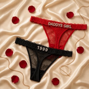 May include: Two pairs of lace thong underwear, one red and one black. The red pair features the text DADDYS GIRL in silver rhinestones on the waistband. The black pair features the number 1999 in silver rhinestones on the waistband. Both have lace trim.