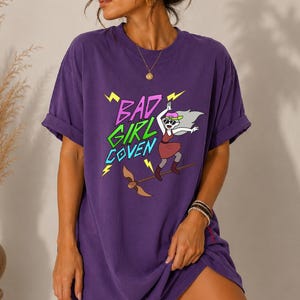 May include: A purple short-sleeve t-shirt featuring a graphic print with the text "BAD GIRL COVEN" in pink and green. The design includes a cartoon character wearing sunglasses and a cap, riding a broomstick, accented by yellow lightning bolts.