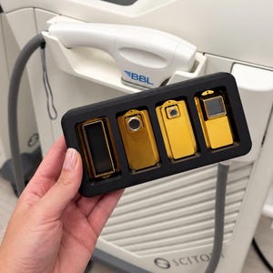May include: A black rectangular tray containing four distinct gold-colored metal attachments for a medical device. The attachments vary in shape and aperture size, featuring rectangular and circular openings. The tray is held in front of a white BBL machine.