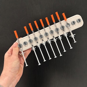 May include: A white plastic organiser tray holding nine clear plastic syringes with orange caps. The tray features circular cutouts for the syringes and is marked with the letters GL, L, S, and C, 2N. Each syringe has a white plunger and a clear barrel.
